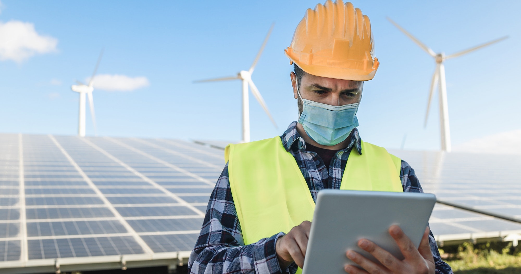 Man worker using digital tablet while wearing safety mask at renewable energy farm